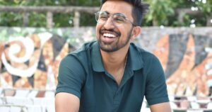 Cheerful young man with glasses smiling outdoors against artistic wall background.