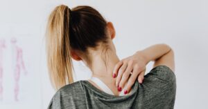 Most People Don't Realize That Your Cellphone Is Changing Your Body, Doctors Say Back view of slim blond female in casual wear with manicure indicating pain spot on back while sitting in front of white wall with paper drawing in clinic