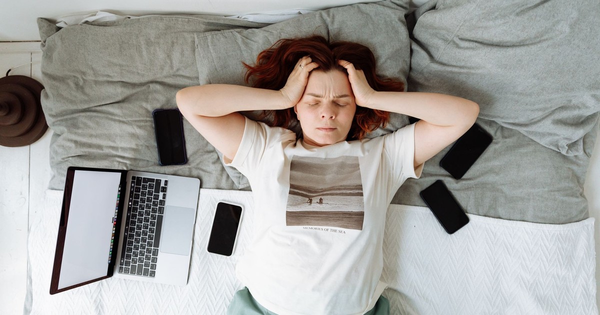 A distressed woman lying in bed surrounded by digital devices, showing signs of stress.