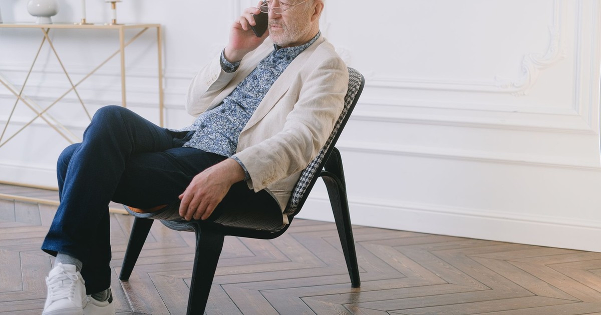 A senior man in stylish casual attire sitting on a chair indoors, using a smartphone.