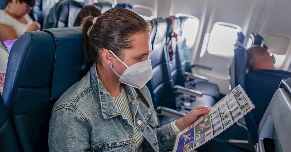 A woman wearing a mask on an airplane seat reading safety instructions.