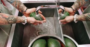 Nearly 1 in 5 Avocados Tests Positive for This — Here's What to Do Colorful tattooed arms washing fresh avocados under a kitchen sink faucet.