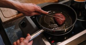 A chef sears a steak in a skillet on an induction cooktop in a professional kitchen.