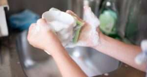 Close-up of hands washing a cup with soap suds in a kitchen sink, representing daily home chores.