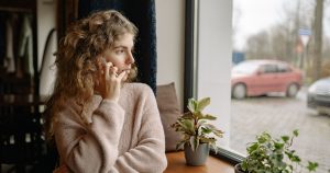 Woman in cozy sweater talking on a phone by the window, surrounded by houseplants, observing outdoors.
