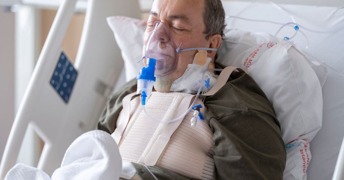 An elderly man lies in a hospital bed wearing an oxygen mask for medical care.