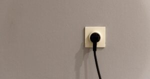 A black plug inserted into a white wall socket on a plain grey wall.