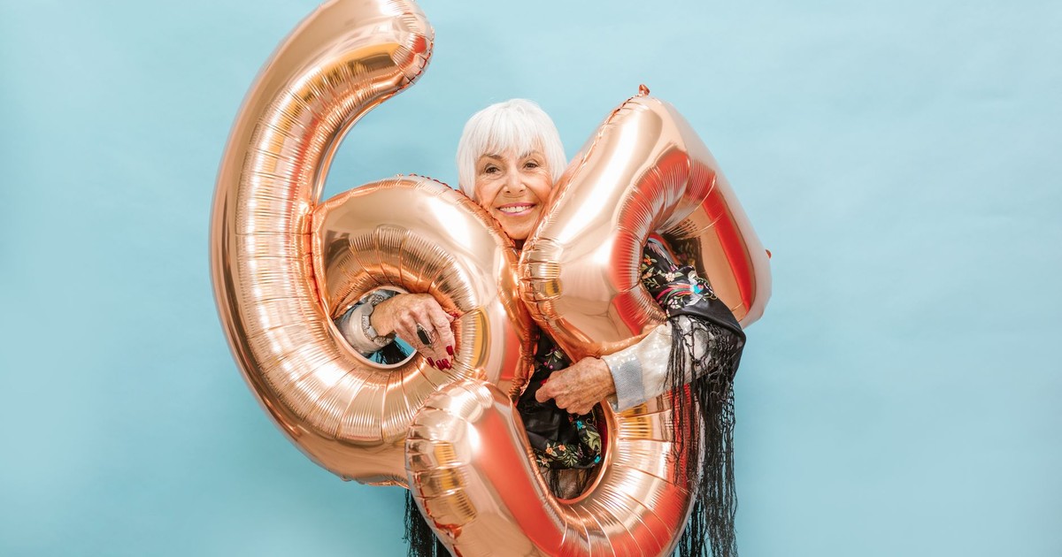 Happy senior woman poses with 64 balloons on a light blue background, exuding joy and celebration.