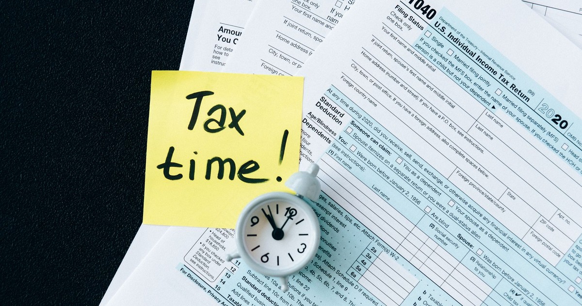 A stack of tax forms with a clock and yellow sticky note saying 'Tax time!' indicating urgency.
