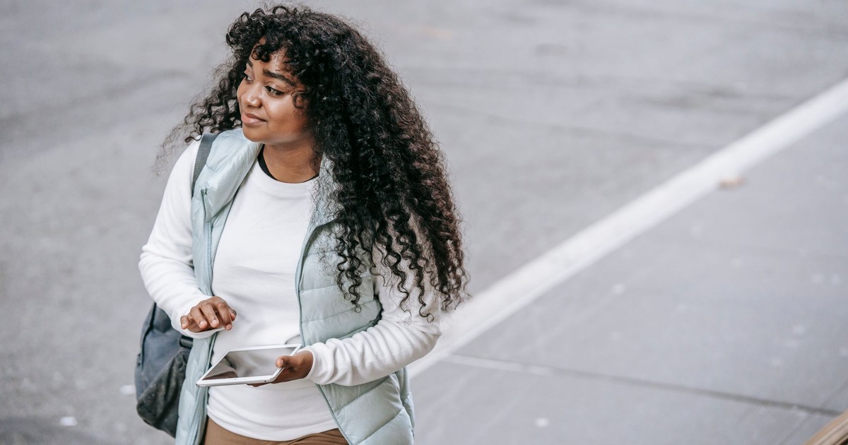 Young black lady wearing casual clothes and backpack while strolling on city street with tablet on walkway near road in daytime and looking away
