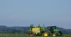The EPA Just Approved 2 Forever Chemical Pesticides on America's Biggest Food Crops Green tractor spraying fertilizer on a vast agricultural field under clear blue sky.