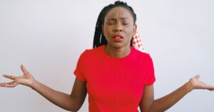 Portrait of a woman in a red shirt expressing confusion with her eyes closed and arms out. Ideal for emotion concepts.