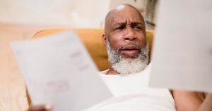 A middle-aged man looks concerned as he reviews household bills, sitting in a comfortable chair indoors.