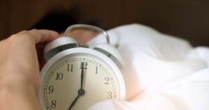 What Permanent Daylight Saving Time Would Actually Do to Your Body A close-up of a hand reaching for a ringing alarm clock, symbolizing waking up in the morning.
