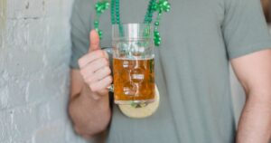 7 Things That Happen to Your Body When You Drink Beer Every Day A person holding a mug of beer, wearing green beads, celebrating St. Patrick's Day indoors.