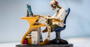 A quirky skeleton figurine wearing headphones sits at a desk with a computer, symbolizing burnout.