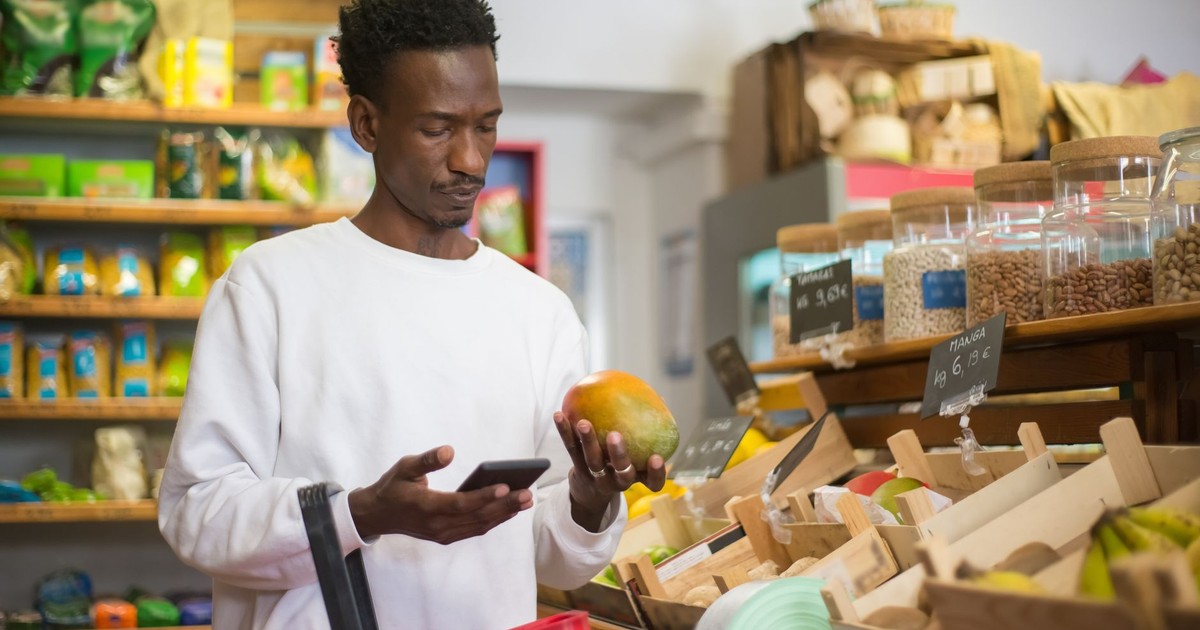 A man examines a mango while shopping with a smartphone in a grocery store.