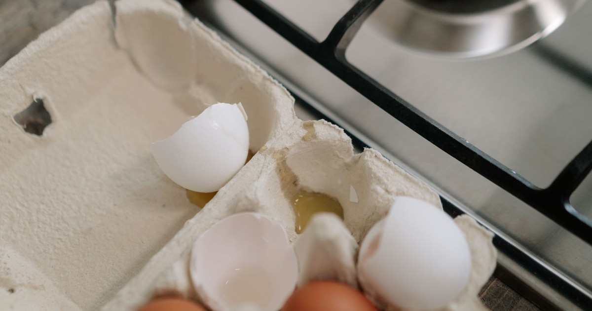 Egg carton with brown and white eggs and broken shells on kitchen stovetop.