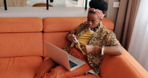 A worried woman sitting on an orange sofa checking her temperature with a thermometer, while a laptop sits open beside her.