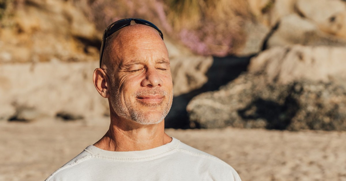 Content senior man with eyes closed, meditating and relaxing on a sunny beach day.