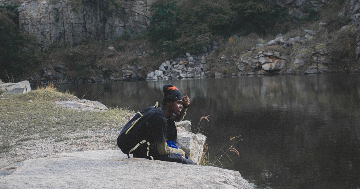 A person sitting alone on rocks, reflecting by a calm lakeshore in nature.