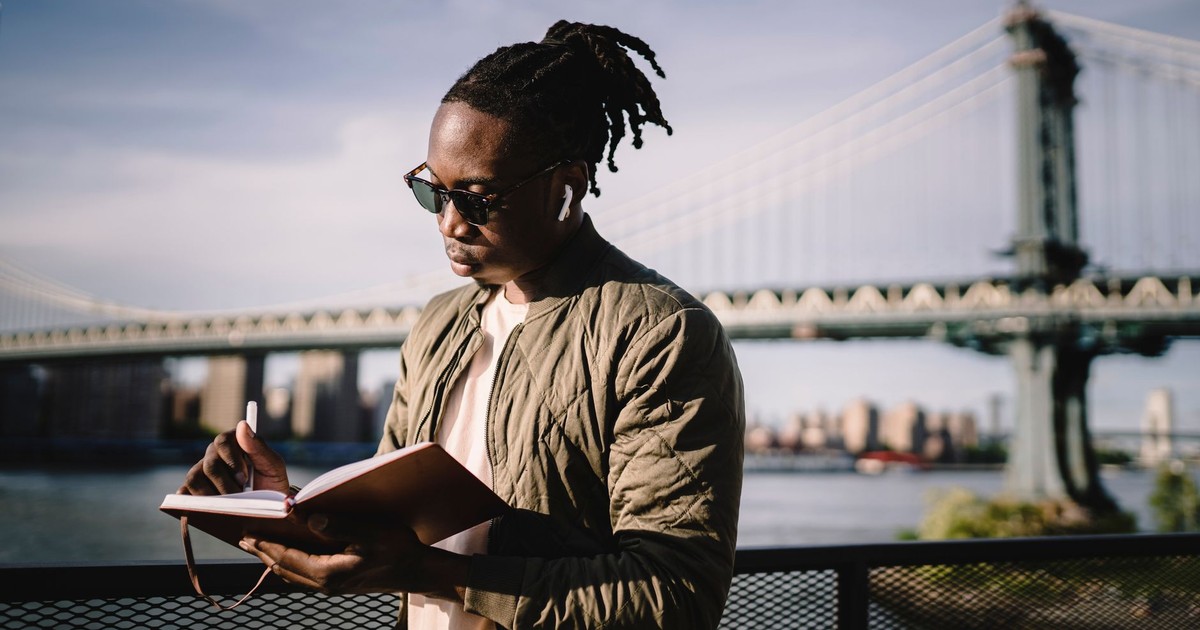 Pensive trendy black guy in sunglasses and true wireless earphones listening to music while writing with pen in open notebook standing on bridge over city river in daylight