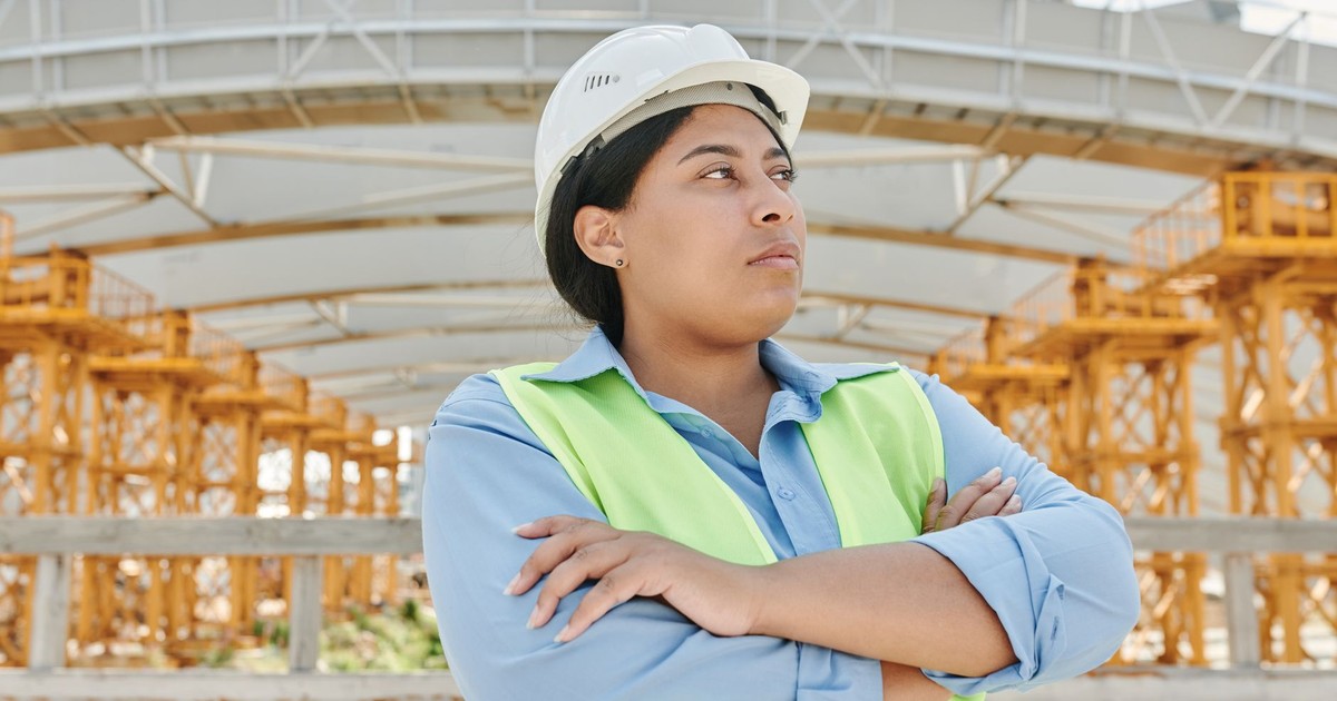 Young female engineer crossing her arms at a construction site, wearing a hardhat and reflective vest.