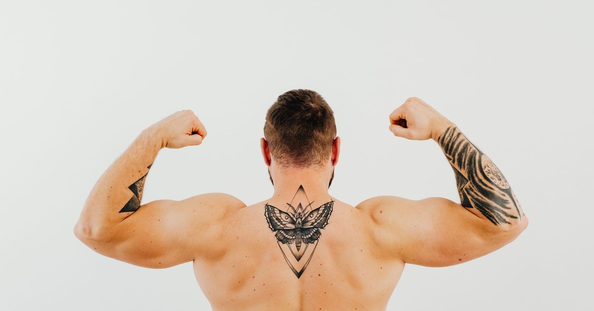 Shirtless man showcasing tattoos and biceps while flexing in a studio setting.