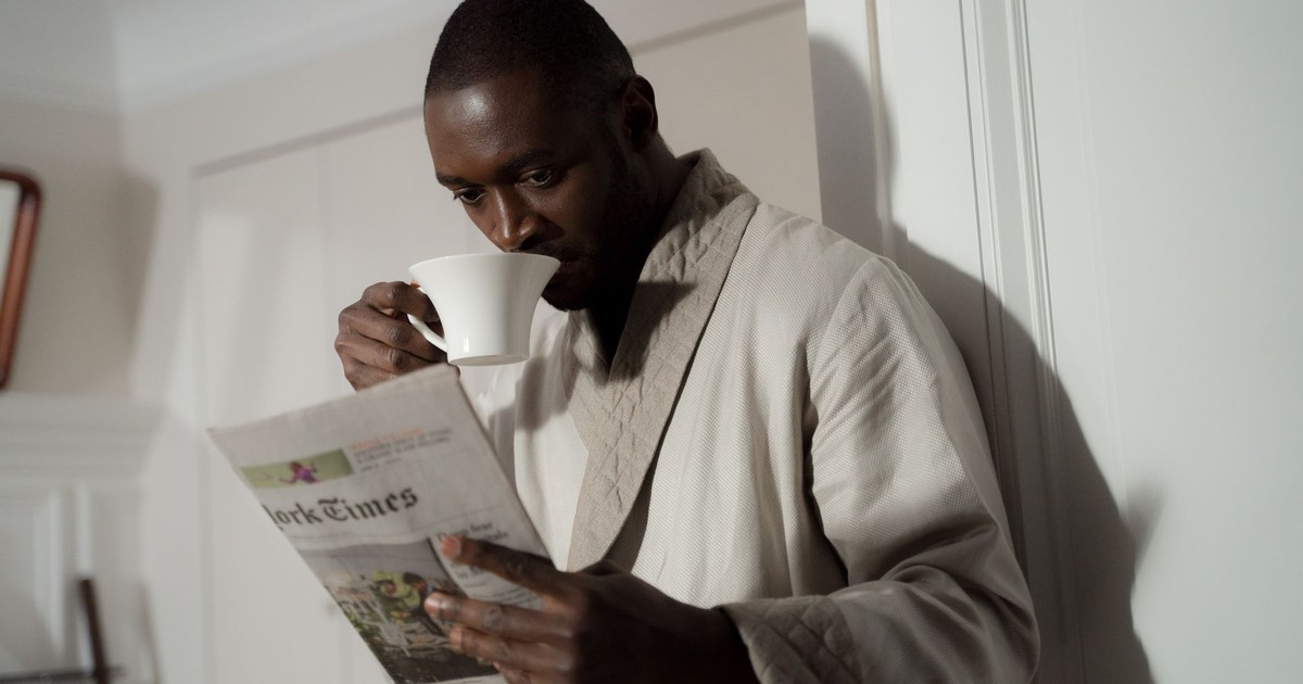 Man in a bathrobe enjoys coffee and reads a newspaper indoors.