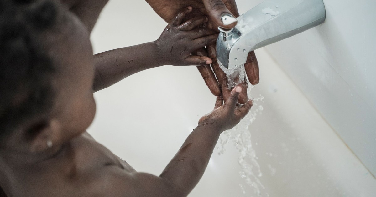 A caring parent assists their young child in washing hands under running water in a bathtub.