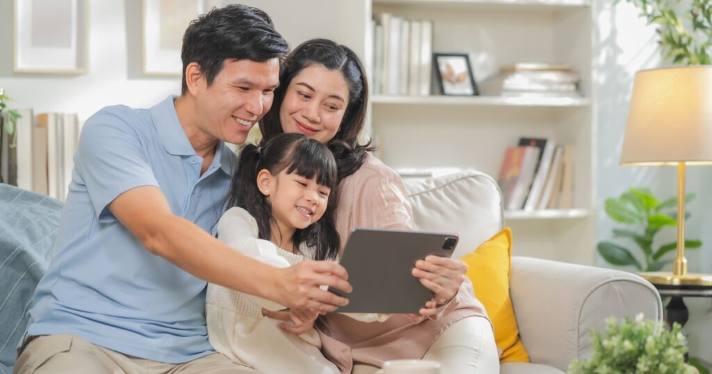 Portrait of happy multi-generation Asian family with dad mom and kids using smartphone digital tablet for video call while sitting on sofa at home during family bonding relaxation time