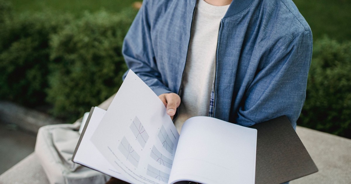 Crop anonymous student in casual clothes sitting on bench in green park and learn project with graphs in daytime