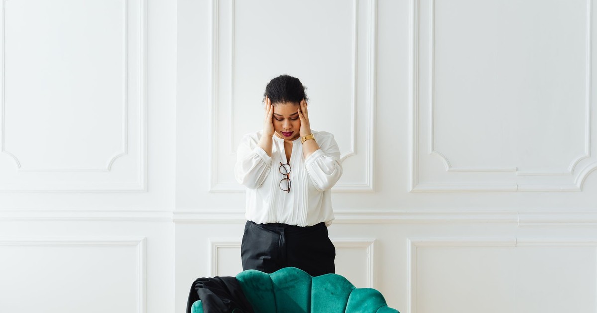 A woman holds her head in stress, standing indoors next to a green chair.