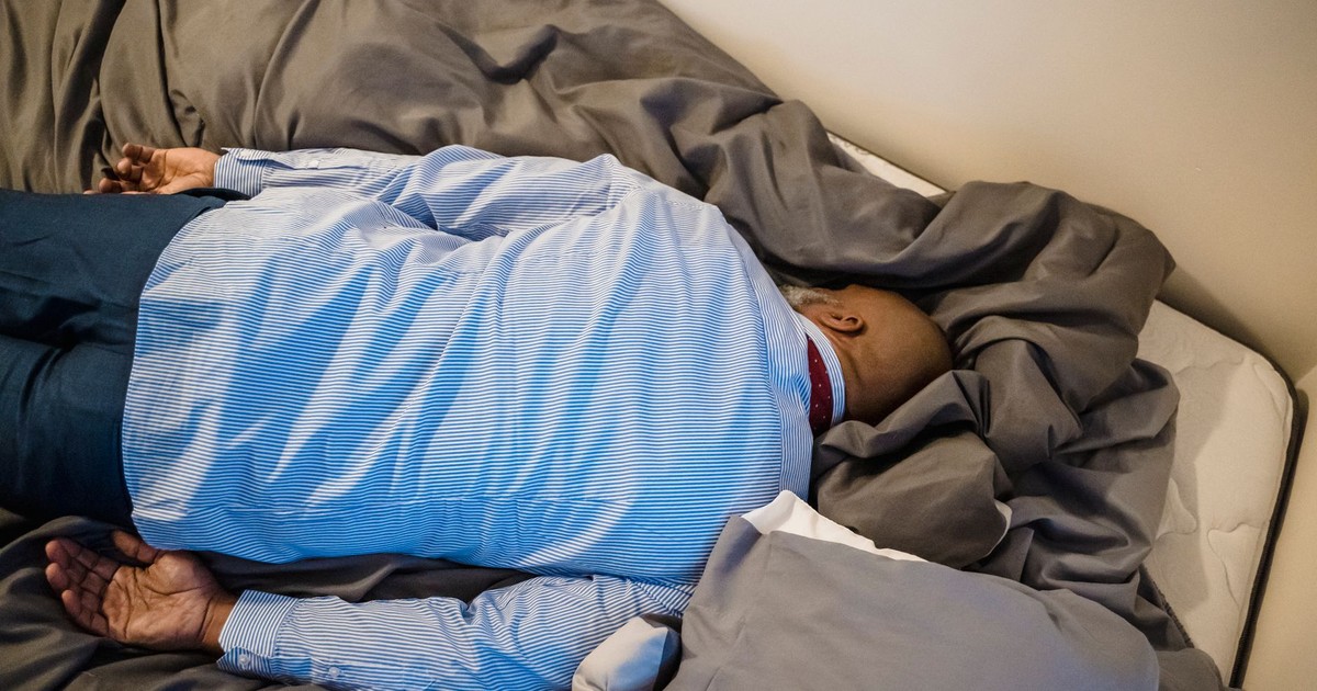 A man in formal attire rests face down on a bed, conveying exhaustion and fatigue.