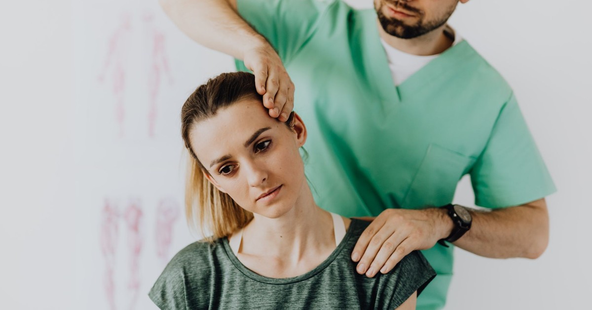 Crop osteopath in uniform and eyeglasses standing with closed eyes while examining neck of young thoughtful female in casual wear in hospital