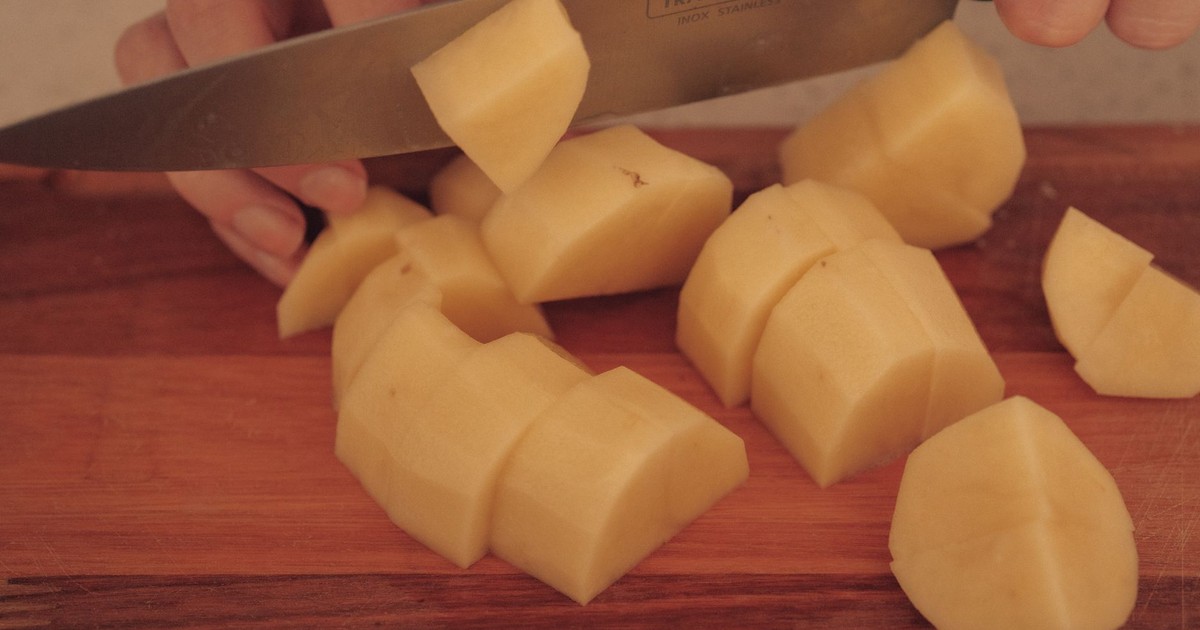 A person cuts potatoes on a wooden board with a kitchen knife.