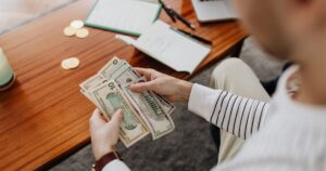 A person counts US dollar bills with a notebook and coins on the desk.
