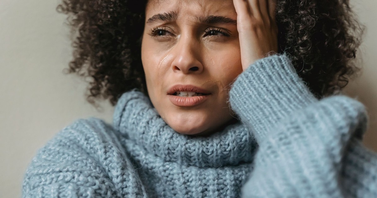 Unhappy African American woman with short curly hair in trendy warm sweater crying touching head and against light background