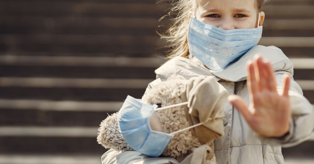 Worried child wearing warm clothes and mask hugging teddy bear in medical mask and showing stop gesture while looking at camera