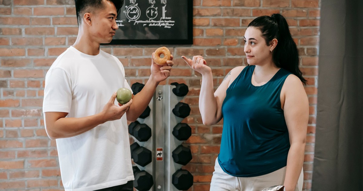 Side view of Asian personal instructor with apple and doughnut offering healthy food to plus size woman while standing in gym