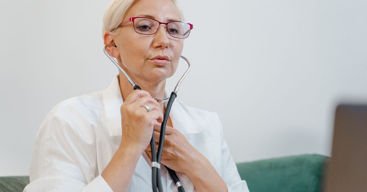 Elderly female doctor using a stethoscope during a virtual consultation in a medical office.
