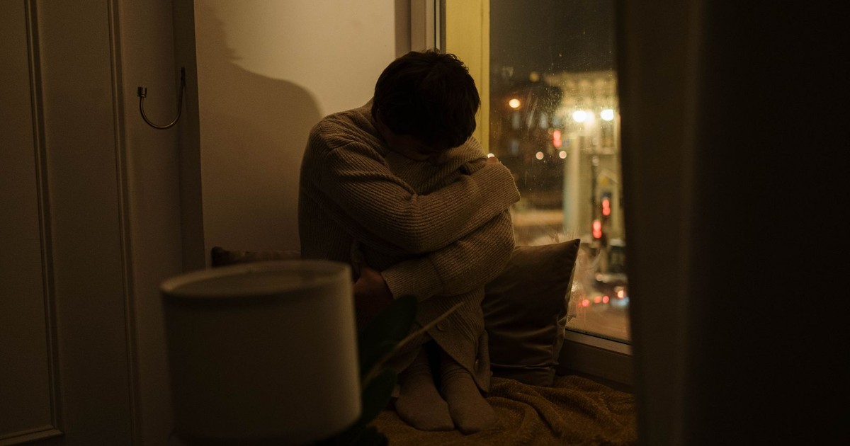 A woman sits alone by a window at night, depicting solitude and reflection.