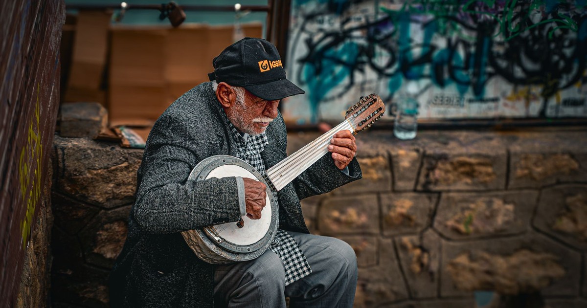 Elderly man in streetwear playing a banjo against graffiti wall. Urban music scene captured outdoors.