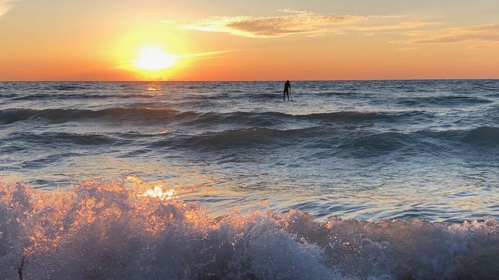bright golden sunset over a wavy body of water with a lone paddleboarder in the distance.