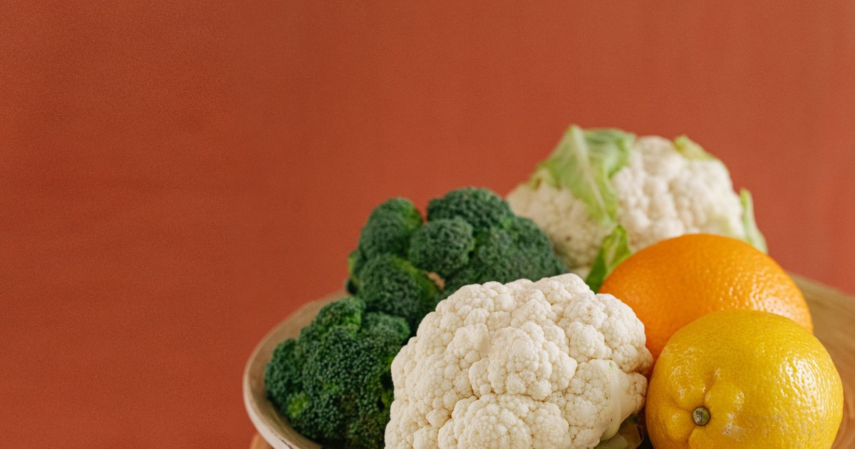 Close-up of fresh broccoli, cauliflower, lemon, and orange on a wooden plate with an earthy background.