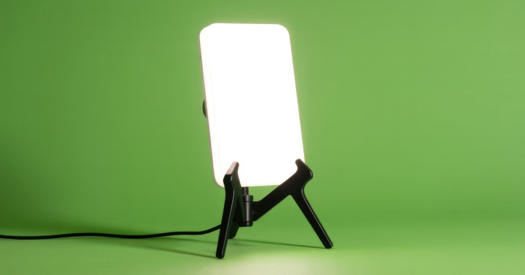 A bright white light therapy lamp with a black tripod stand is prominently displayed against a vibrant, solid green background, with a power cord visible.